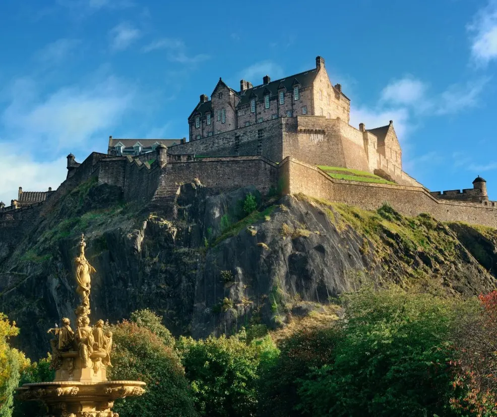 Book-Inspired Places, Scotland travel, Edinburgh, historic castle, fortress, medieval architecture, European heritage, city viewpoint, dramatic landscape, and historical tourism are vividly represented in this mid-to-low-angle photograph of the majestic Edinburgh Castle, which stands powerfully atop the rugged volcanic Castle Rock, contrasting with the lush green foliage and the ornate Ross Fountain in the foreground under a bright blue, cloudy sky. Book your unforgettable Scottish adventure with Saar Holidays.