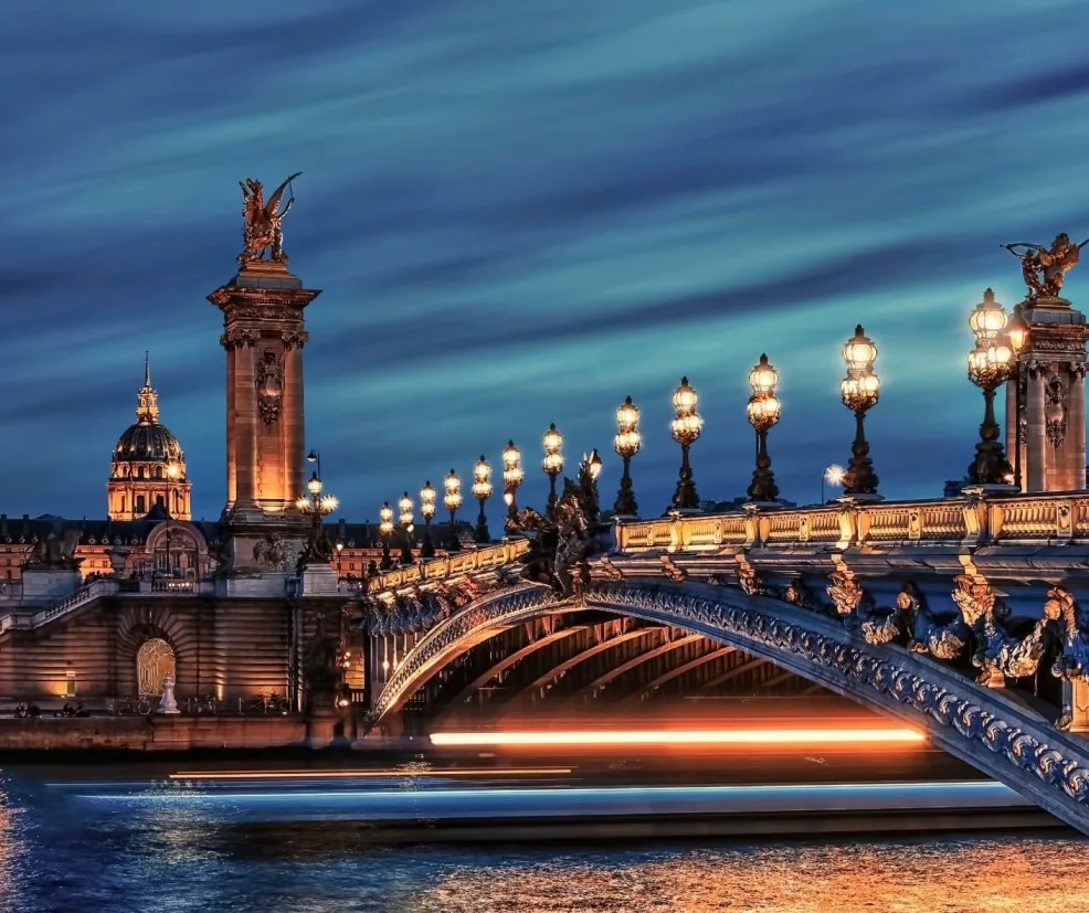 Book-Inspired Places, Paris travel, iconic bridge, Europe vacation, romantic getaway, Pont Alexandre III at dusk, France tourism, architectural marvel, historical landmark, and European city break perfectly capture this dramatic, long-exposure night shot of the illuminated Pont Alexandre III and the dome of Les Invalides against a deep blue twilight sky, with the golden reflections creating light trails on the Seine River. Book your next Parisian night tour with Saar Holidays.